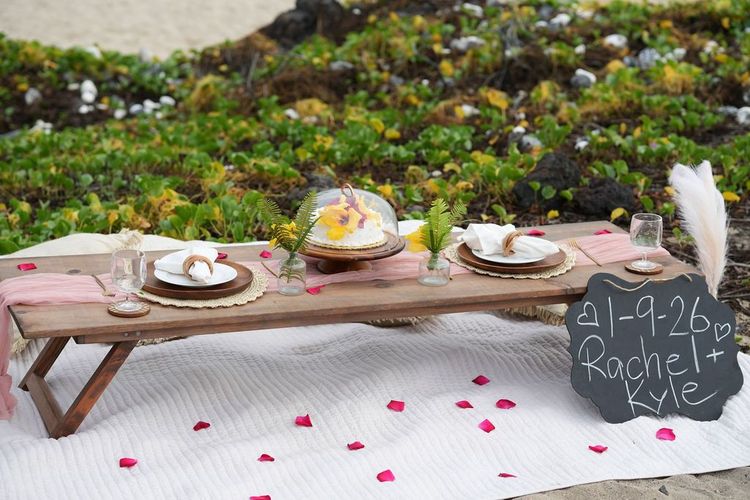 Elegant beach picnic setup with white umbrella, cushions, and scattered rose petals on Hawaii coastline