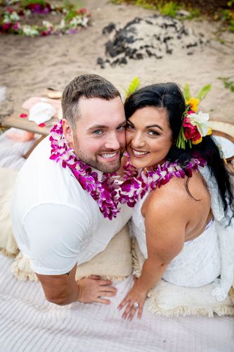 Newlywed couple lying on sandy beach wearing vibrant pink plumeria flower leis in Hawaii