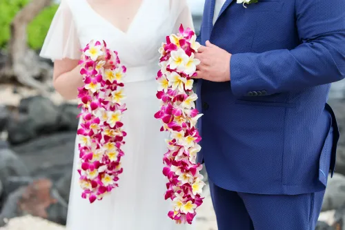Groom in navy blue suit holding two vibrant pink and purple flower leis for Hawaii ceremony