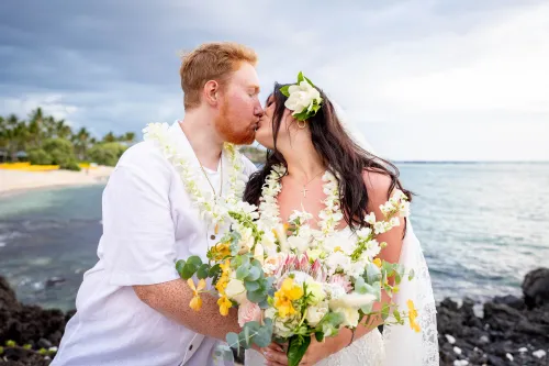 Newlywed couple with tropical yellow bouquet standing on black volcanic lava rocks by ocean in Hawaii